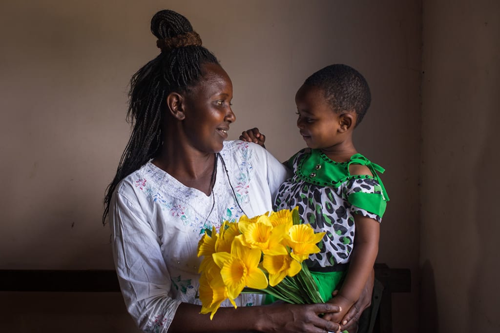 Winner, a Ugandan mother holds her infant son, Godnester and yellow bouquet of flowers. They are smiling at each other.